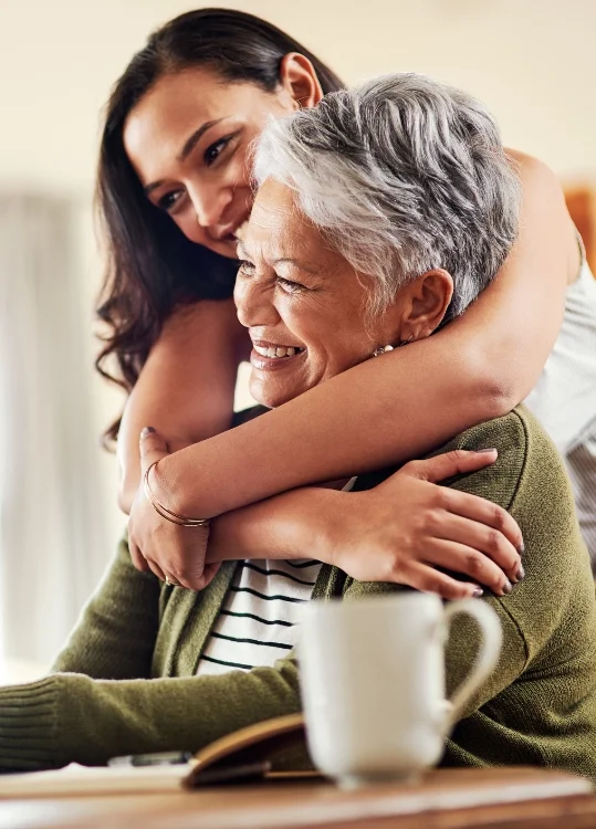 adult daughter leans over the back of her grandmother's chair to hug her from behind, both smiling