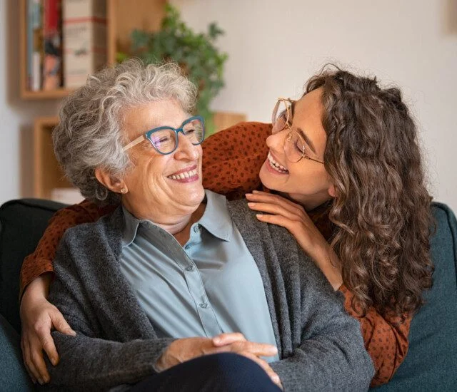 adult daughter leans over the back of her grandmother's chair to hug her from behind, both smiling