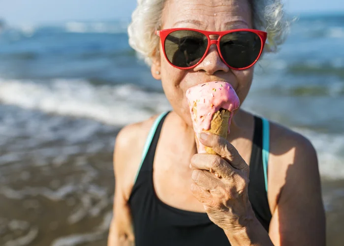 senior woman in bathing suit and red sunglasses eats ice cream cone at the beach