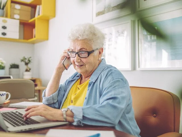 senior woman sits in her kitchen at her laptop, browsing while talking on the phone