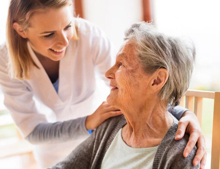 seated senior woman smiles up at her female caregiver, who places her hands gently on her shoulders