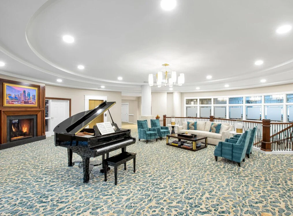 elegant second-floor lounge with seating, fireplace, and grand piano at Claridge Court Senior Living Community