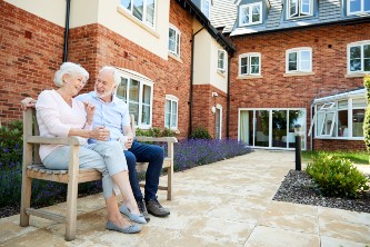 senior couple sit on bench laughing and conversing outside of their senior living community