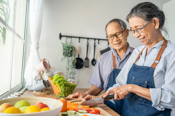 senior couple wearing matching aprons smile while prepping vegetables for a meal in their kitchen