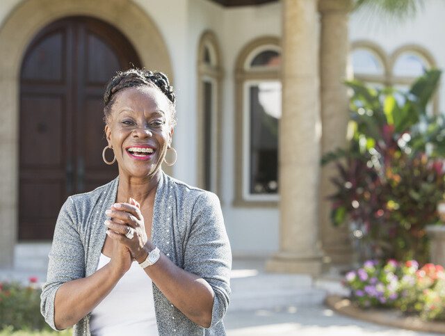 senior African American woman smiles and clasps her hands together while standing in front of her stunning and lavish home entrance