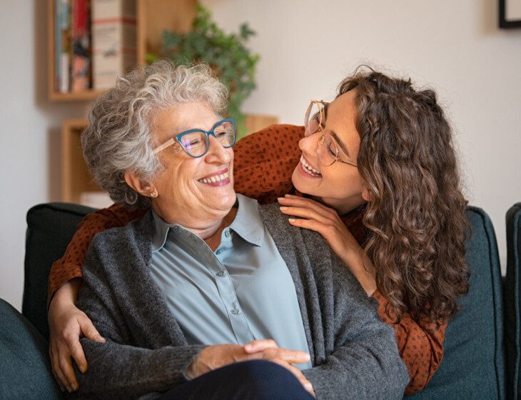 adult daughter leans over the back of her grandmother's chair to hug her from behind, both smiling