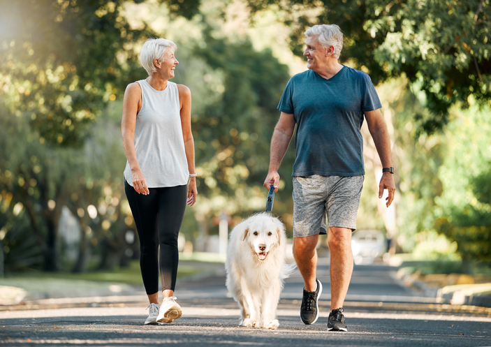 senior couple in athletic clothing smiles and walks their dog on a scenic path