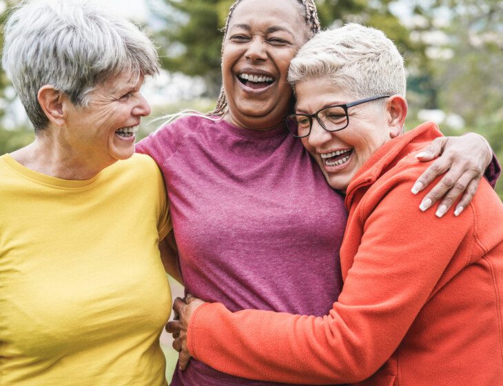 three close senior female friends in athletic clothing laugh and hug outdoors