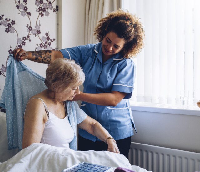 caregiver smiles while helping a senior woman get dressed in her home