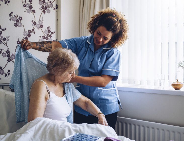 caregiver smiles while helping a senior woman get dressed in her home