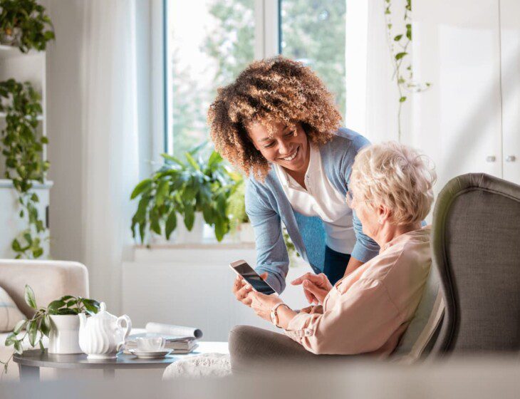 seated senior woman smiles up at her female caregiver, who's helping her navigate her smart phone