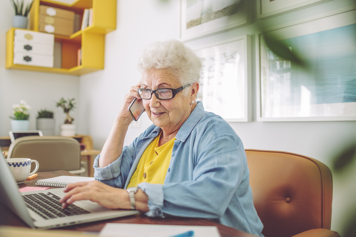 senior woman sits in her kitchen at her laptop, browsing while talking on the phone