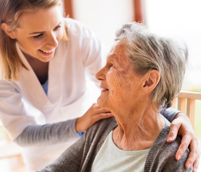 seated senior woman smiles up at her female caregiver, who places her hands gently on her shoulders