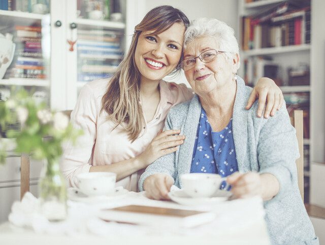 senior woman and her adult daughter smile and lean close for a picture while seated and having tea