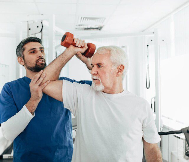 senior man holds dumbbell in one arm with the help of occupational therapist