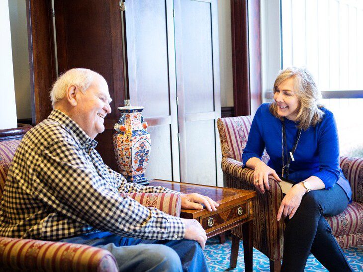 male and female senior friends smile and converse, seated in chairs next to one another in a lounge