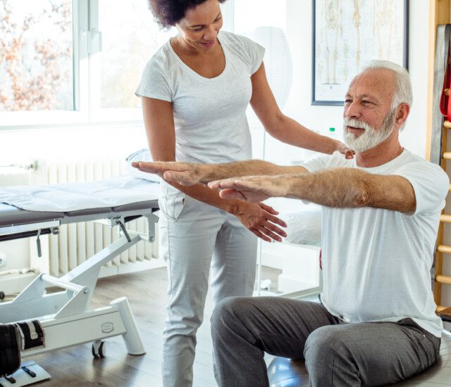 senior man works on mobility while seated on an exercise ball, guided by a physical therapist