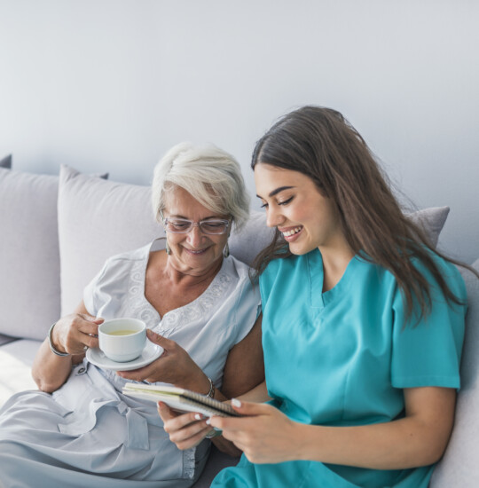 Senior woman smiles and drinks tea on the couch while spending time with her caregiver