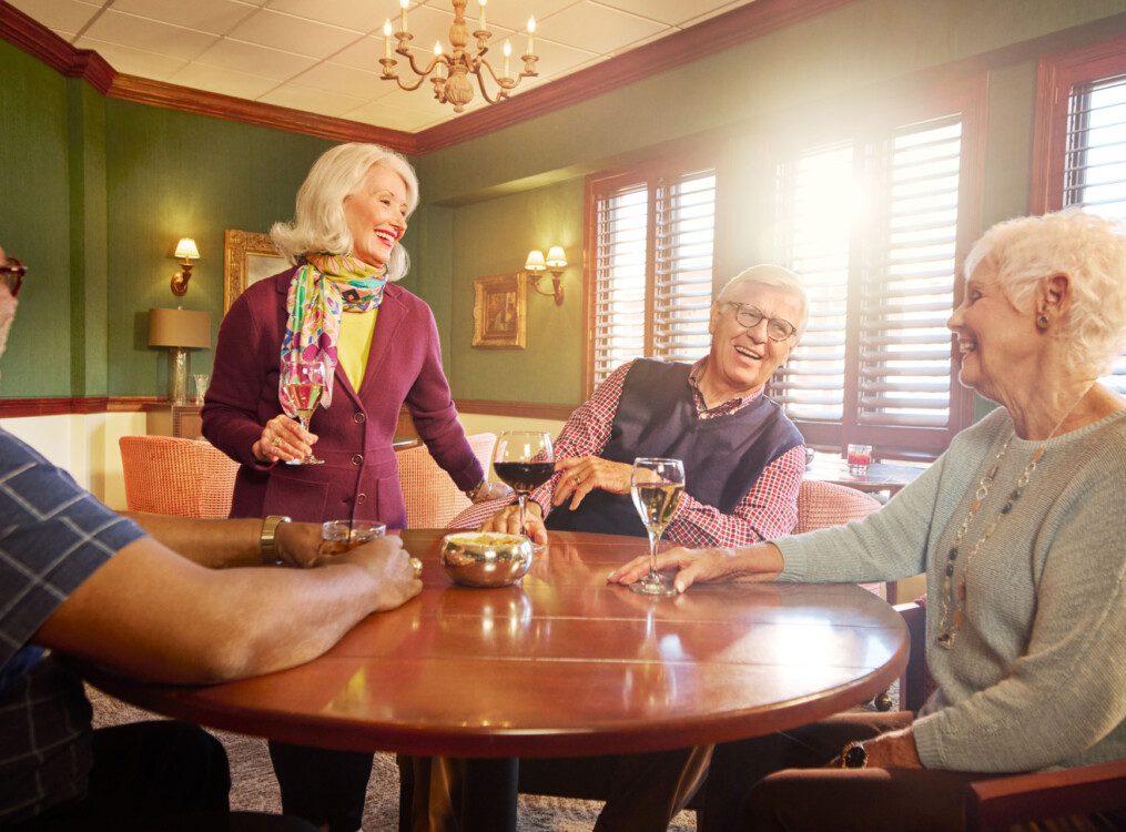 group of senior friends sit at a circular table, smiling and drinking wine in the pub at Claridge Court Senior Living Community