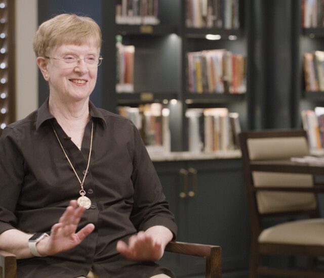 Senior woman in black button-up shirt smiles and sits in chair for interview